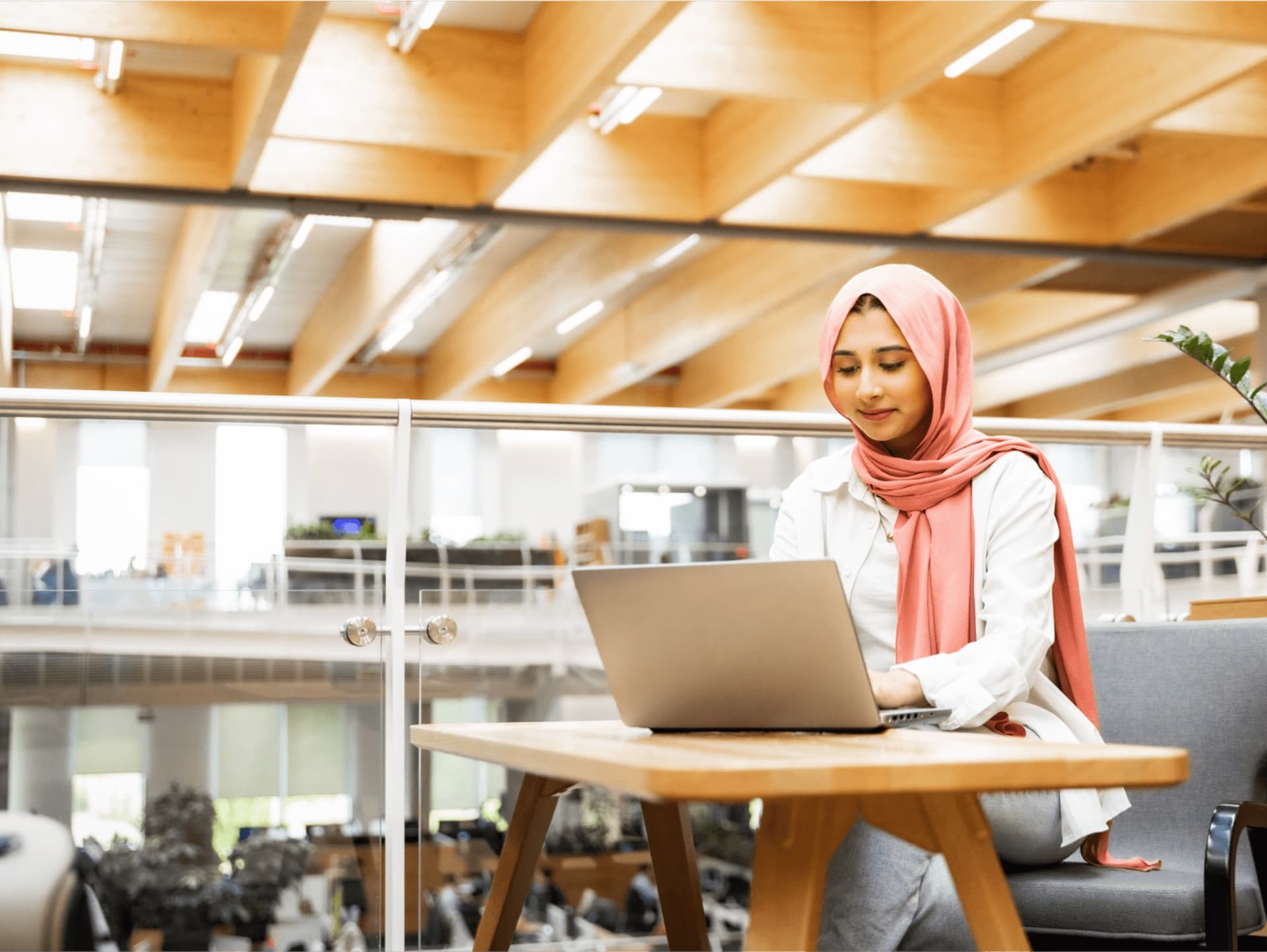 A woman sitting in an open indoor space looking at and typing on an open laptop