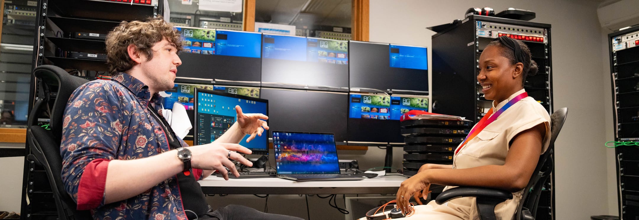 Two team members chat in a control room with screens and laptops around them, one gesturing while the other listens and smiles.