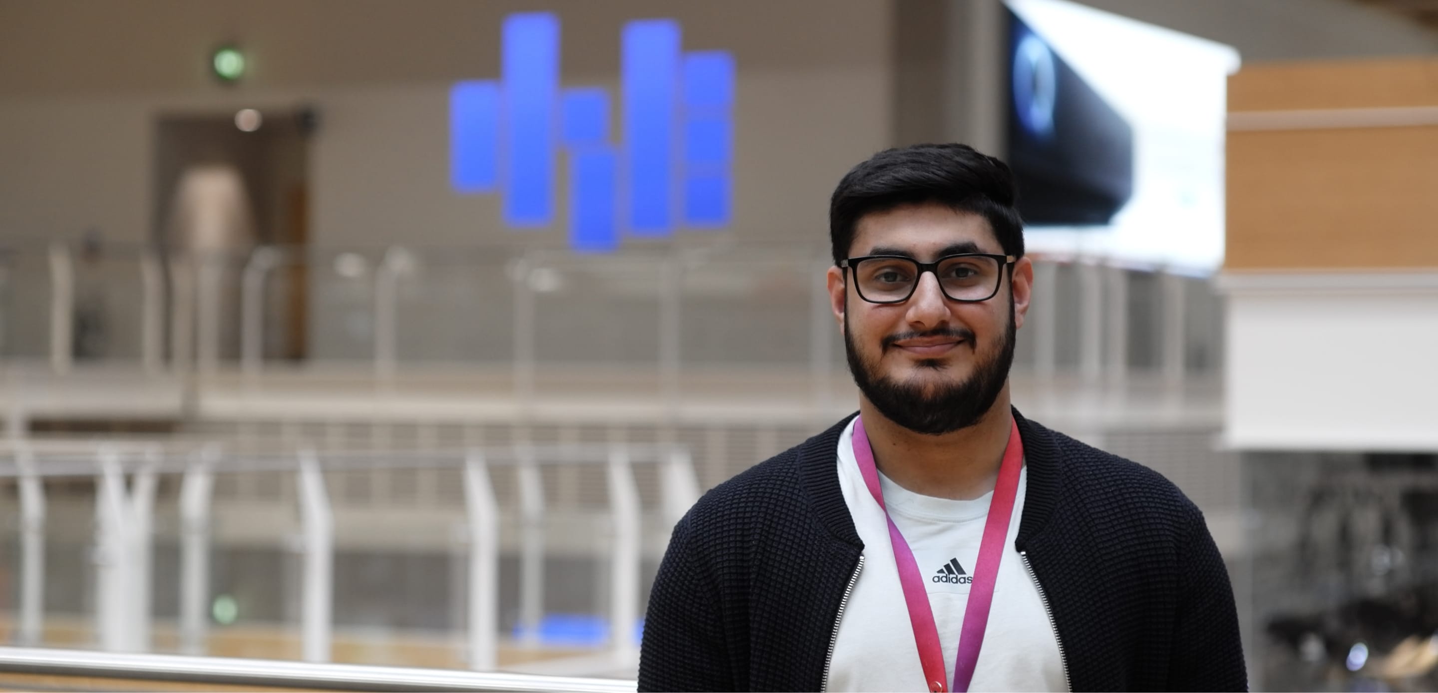 A man in glasses and a dark cardigan smiles in an indoor office area, wearing a Sky lanyard and a white Adidas t-shirt.