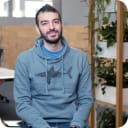 Head shot of man in a blue jumper with a shark print, sat in front of a white shelf with green plans