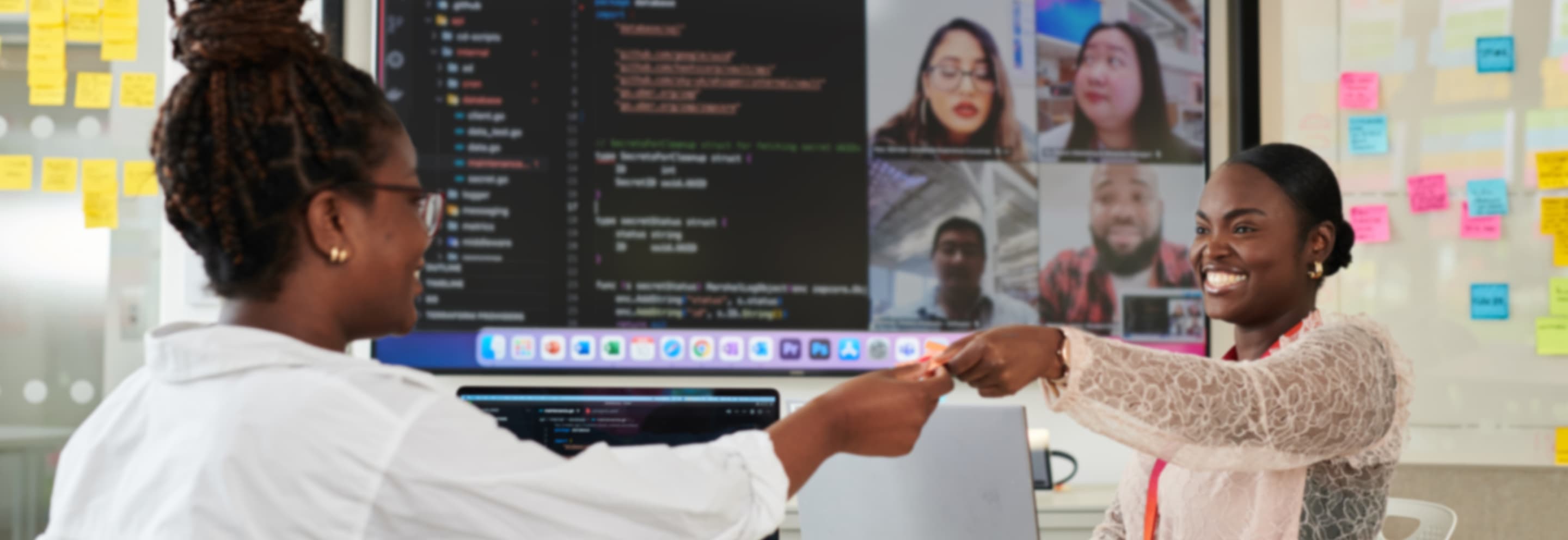 Two women smiling and fist bumping in an office, with a screen showing a video call and coding interface in the background.