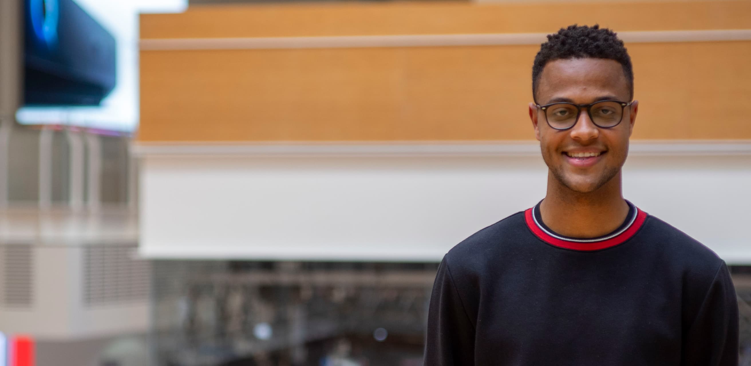 A smiling man in glasses and a dark sweatshirt stands indoors near a balcony, with a blurred tech-themed backdrop behind him.