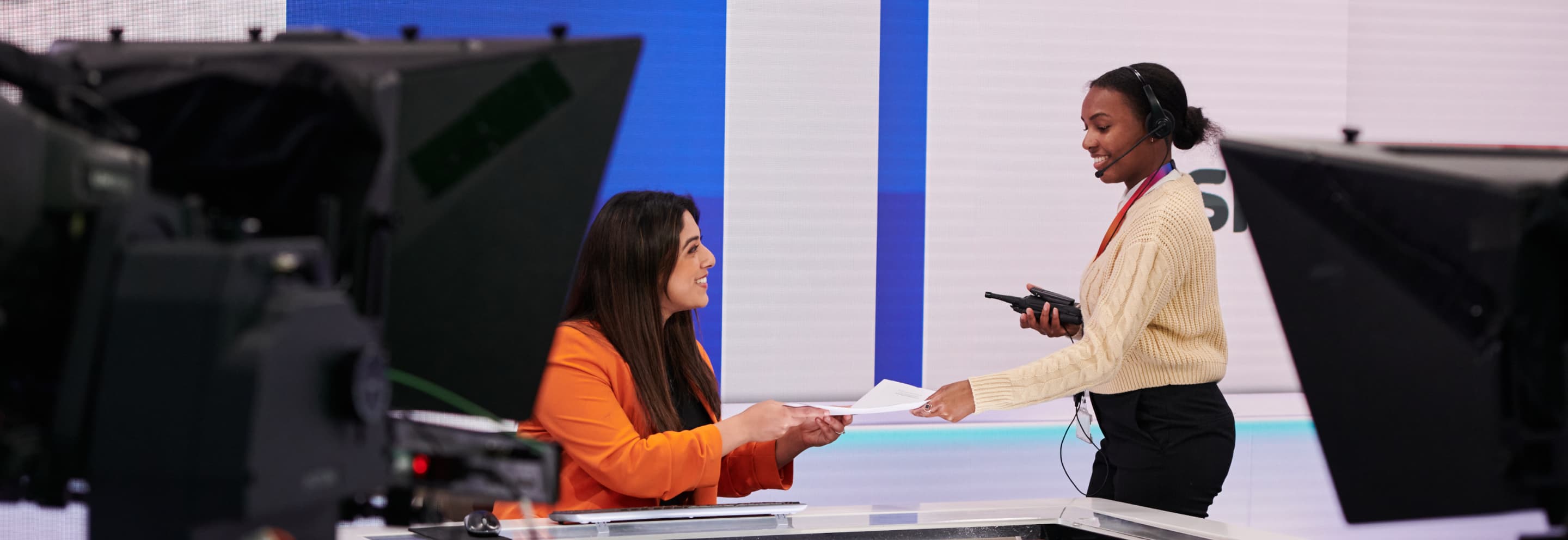 young woman handing a female news presenter some papers