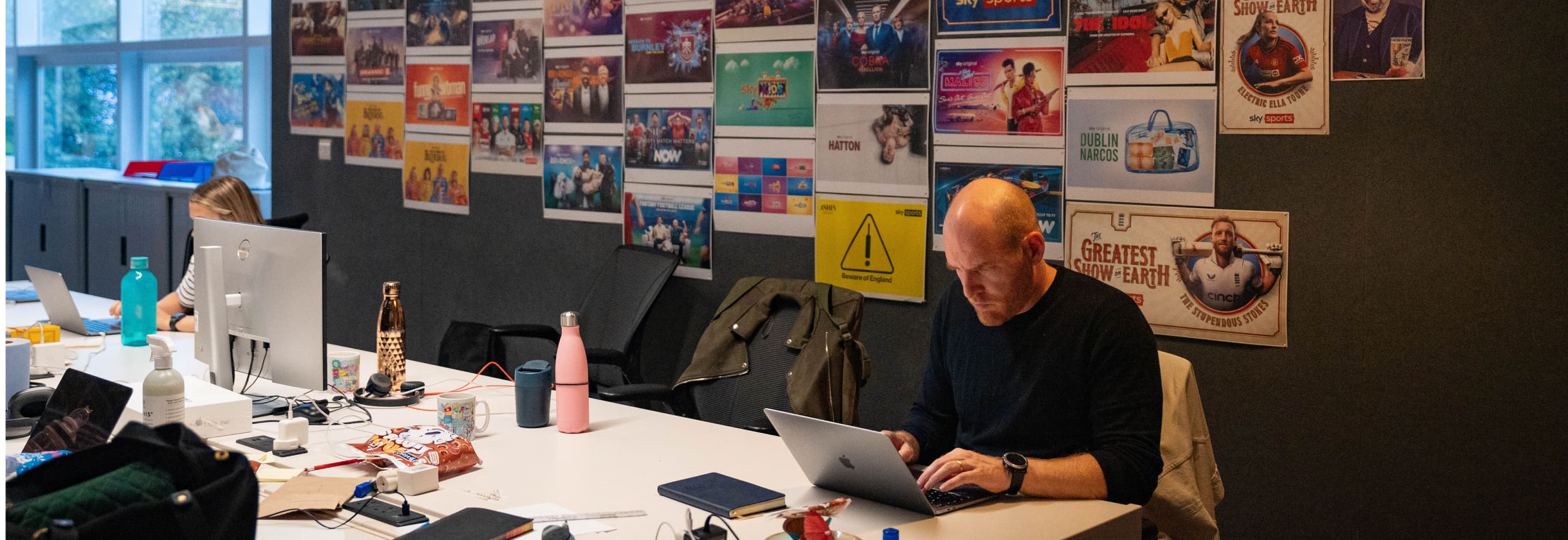 Man working on laptop in creative office, walls covered with TV and film posters, desk cluttered with bottles and snacks.