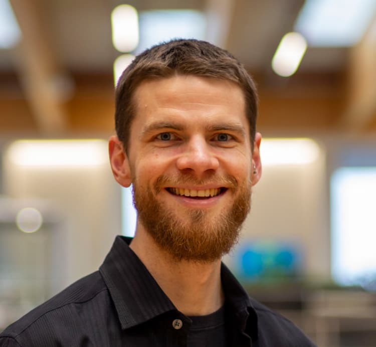 Man with short beard and dark shirt smiling in a modern office setting with soft background lighting.