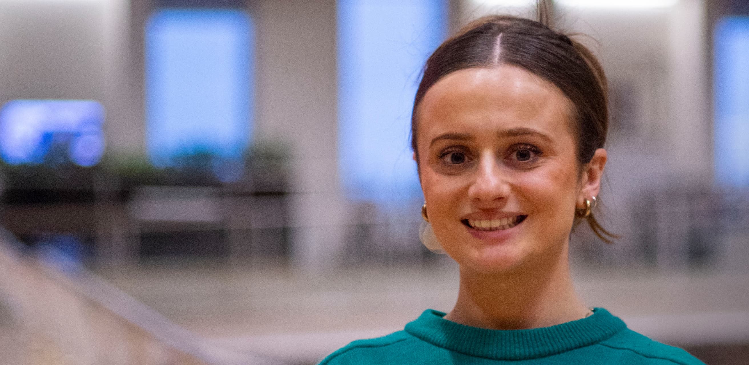 Woman in green jumper smiling at camera in softly lit office with blurred blue and white lights behind.