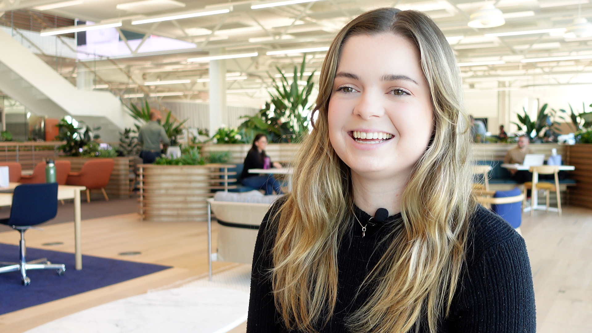 Caroline preparing for a video interview, behind her is a busy Sky Innovation Centre open area, where colleague discuss work over coffee tables.