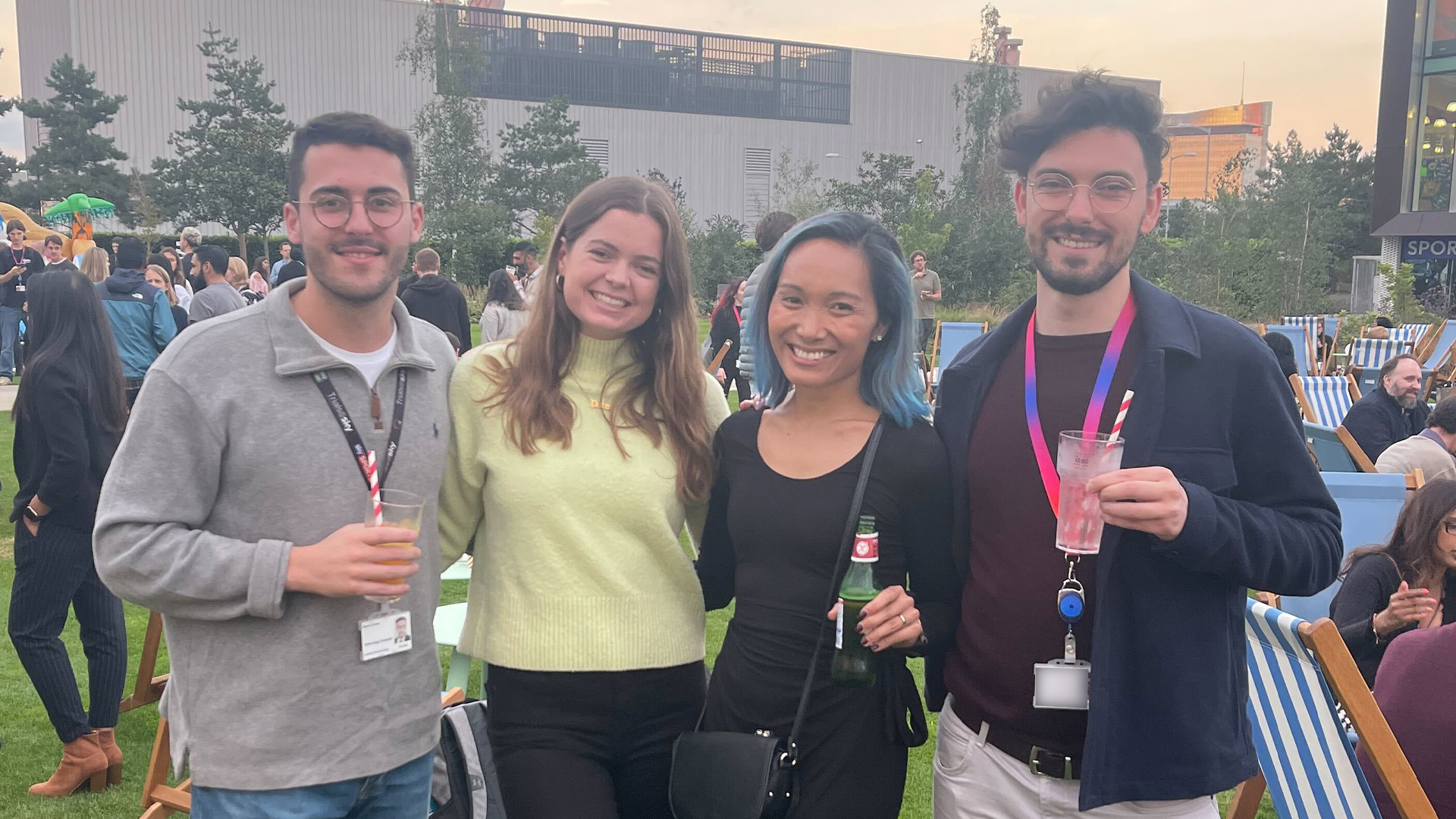 Caroline posing with colleagues for a team photo, during an after-work drinks reception. The sun sets in the background, as colleague make use of the green space outside of Sky Central, chilling out on the nearby deck chairs.