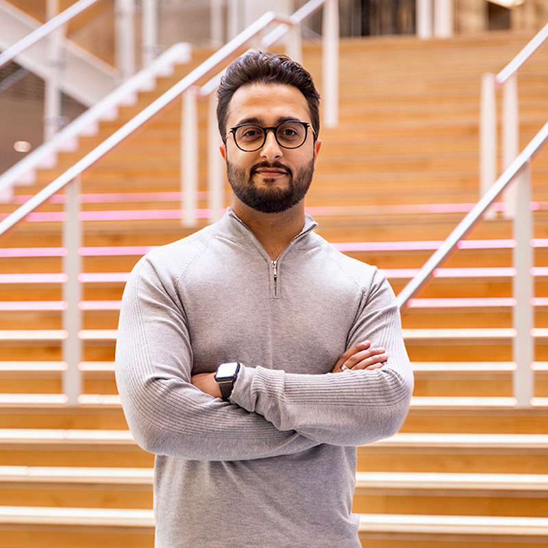 Person in grey top and glasses stands with arms crossed on indoor staircase with pink lights.
