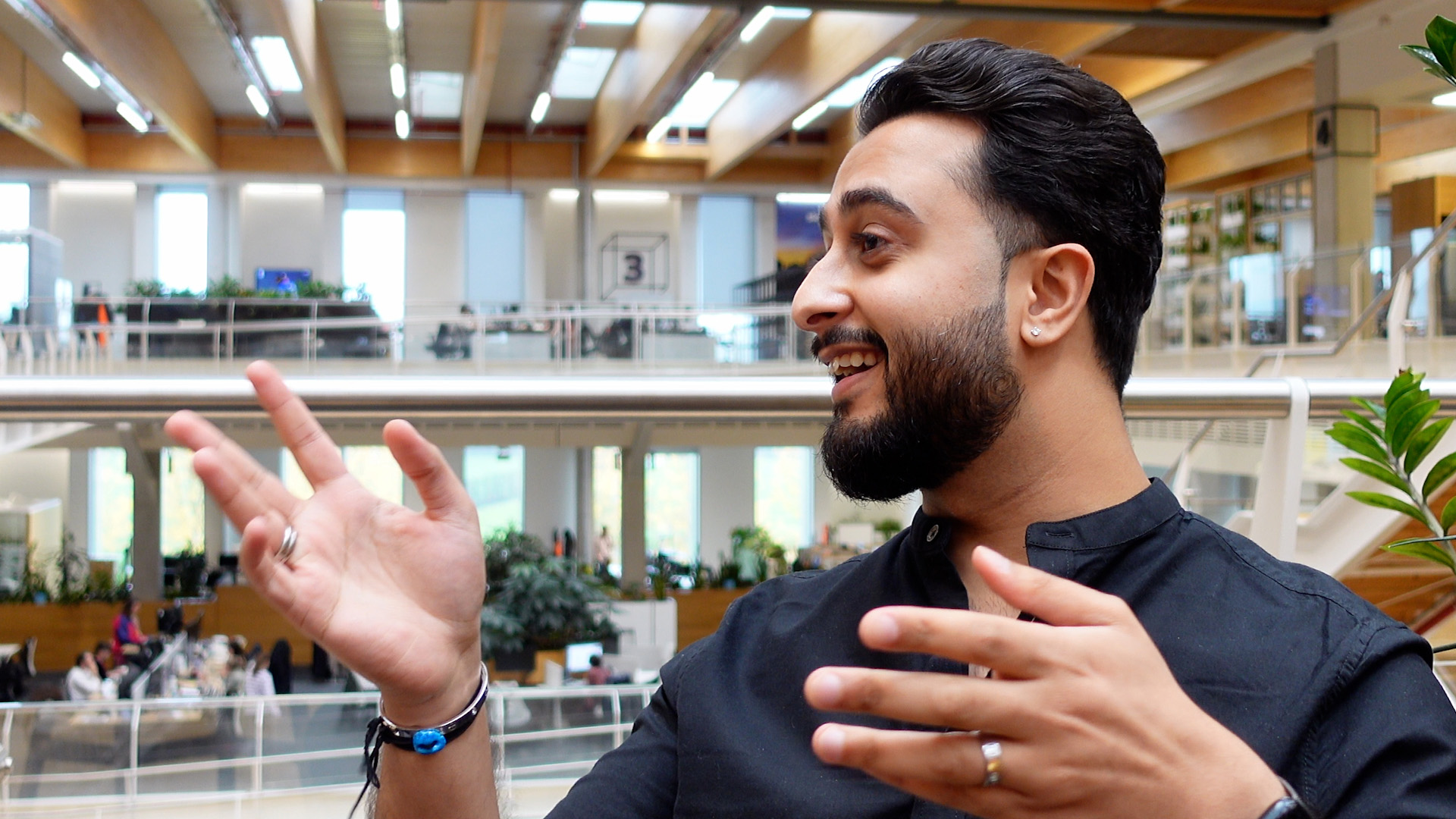 Aman speaking to a colleague off to the left of the camera. Behind Aman is a large open area, within which floors one and two of Sky Central can be seen, with colleagues walking to their desks.