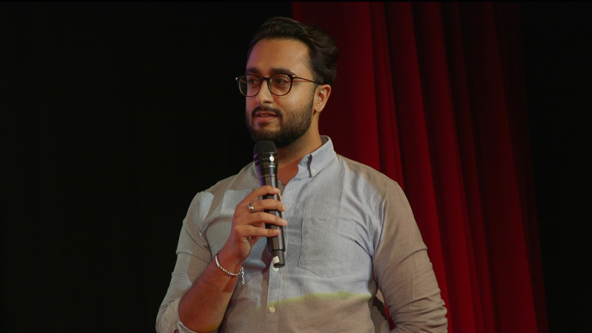 Aman delivering a speech to graduates in the Sky Cinema in Osterley. Aman is holding a microphone, as a red velvet curtain drapes in the background.