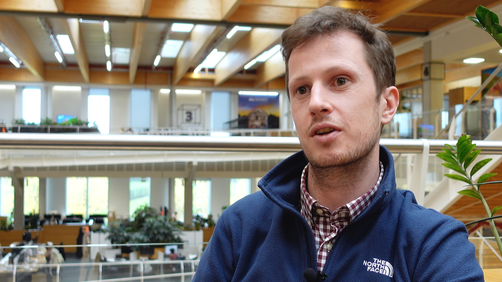 Rob answering a question given to him by the interviewer, who sits behind the camera. Behind Rob is an expansive view of the Sky Central office, as colleagues in the distance are walking past beneath large ceiling windows.