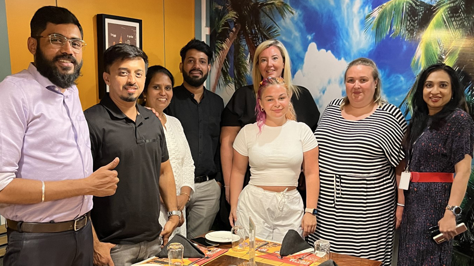 Bekky and her team pose for a team photo. A dining table with empty plates is in the foreground, while the back wall displays a vibrant image of blue skies and palm trees.