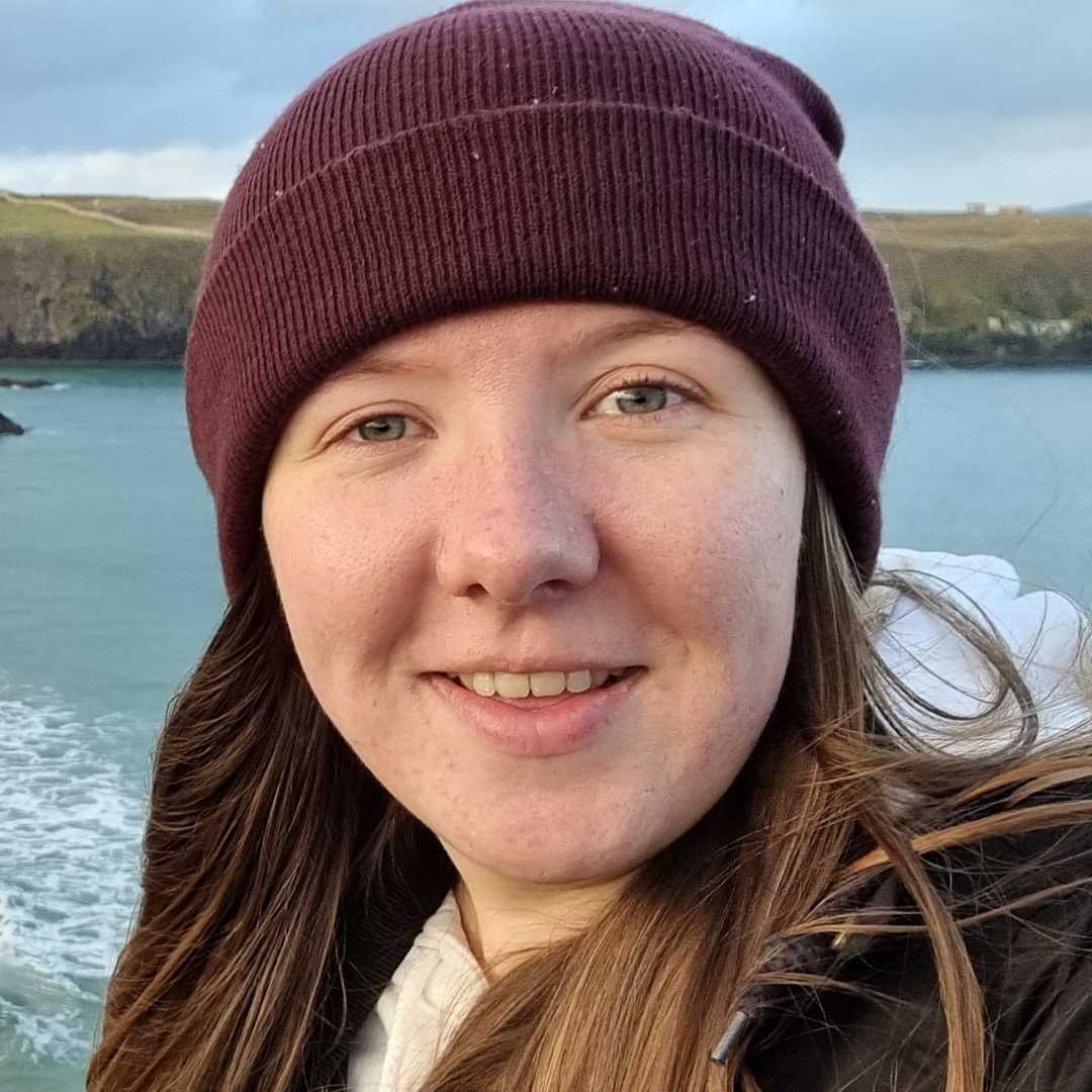 Lizzie smiling in a purple hat stood by the coast