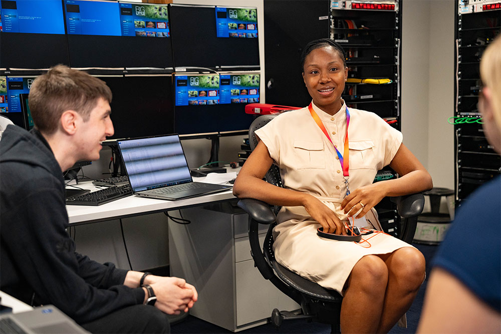 Malissa looking off to the right of the image towards a colleague. Malissa is sat down in a desk chair in front of an array of mounted screens, and a laptop on the desk. A colleague leans in from the left as Malissa discusses work with the team.