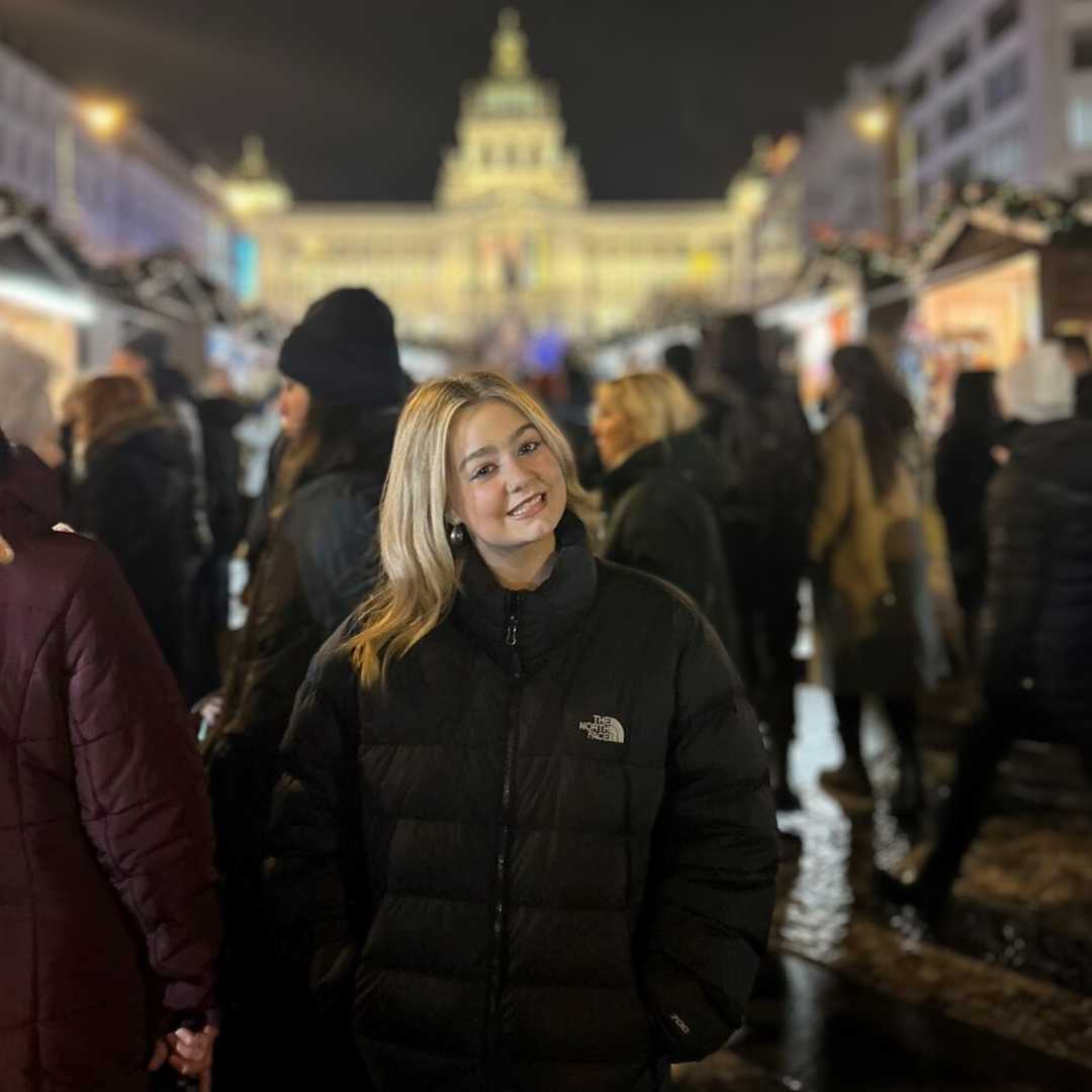 Woman in a black puffer jacket at a festive outdoor market at night, with blurred people and a lit building in the background.