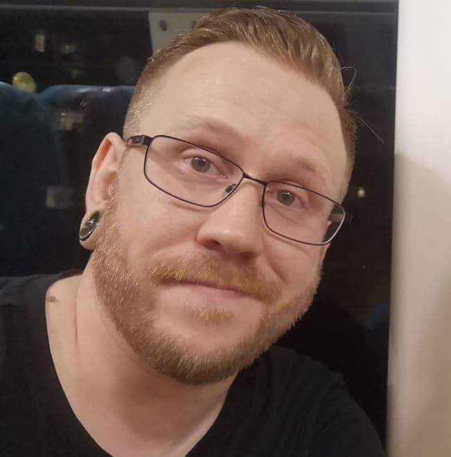 Joe has his photo taken on the window seat of a train. Outside the window is dark, in which the reflection of the other train seats can be seen.