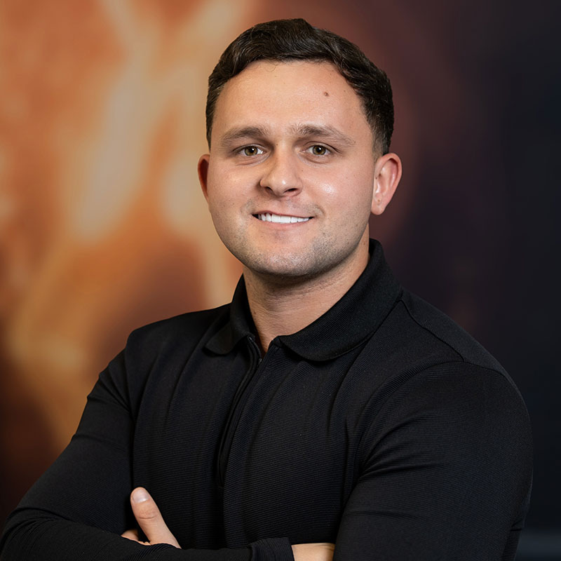 Man in a black polo shirt smiling with arms crossed, standing against a dark orange blurred backdrop.