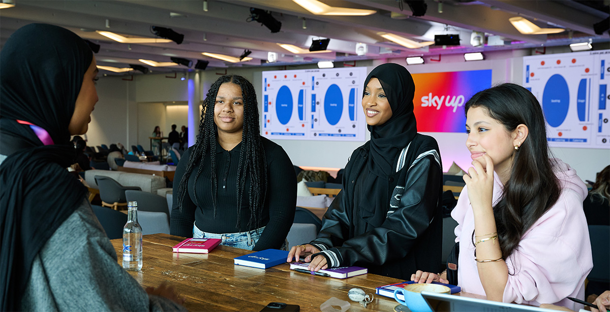 Salma, and other Sky colleagues, speaking over a wooden table with Sky-branded notebooks placed near them. In the background is the 'SkyUp' logo, placed in front of the Sky spectrum, flowing from bright orange to navy blue - neighboured by two slide decks displaying various graphs and data.