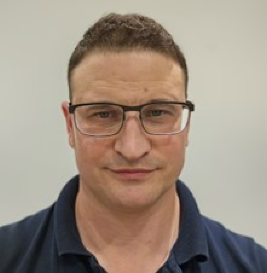 Close-up headshot of man wearing black polo shirt and glasses against a plain background.