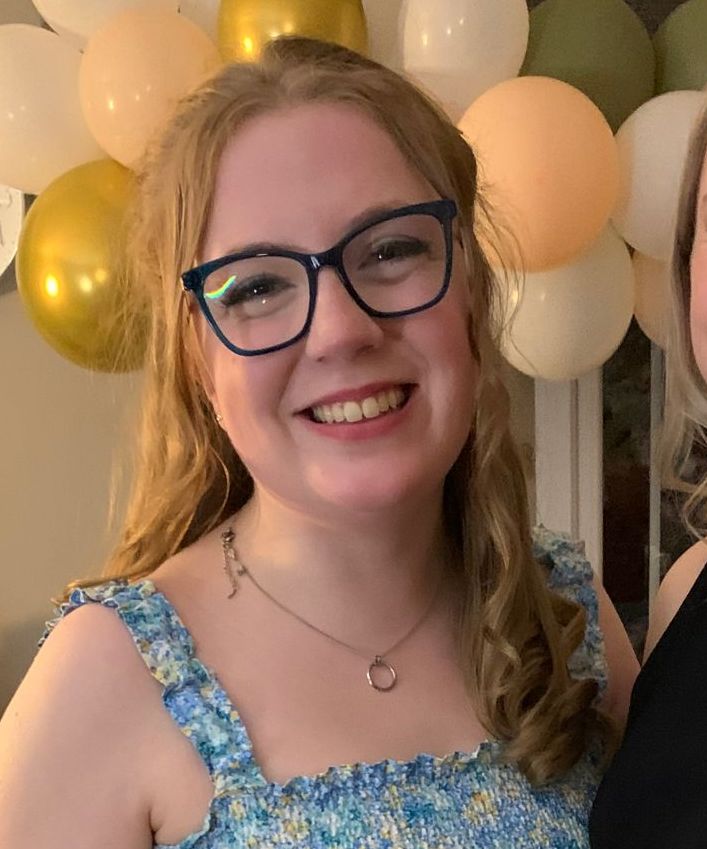 Woman with glasses smiling in front of gold and white balloons, wearing a blue floral dress.