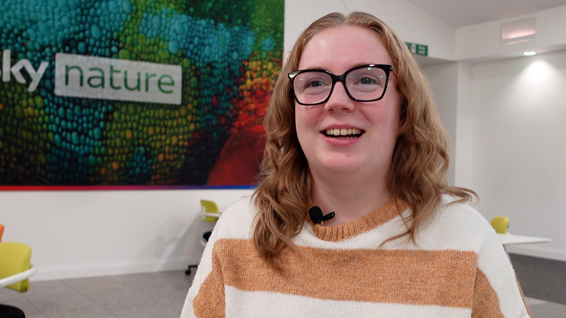 Carys is looking to the left of the camera, smiling towards the interview. Behind Carys is a Sky Nature logo layered above an image of a chamelon. There are office chairs and desk spaces directly behind Carys.