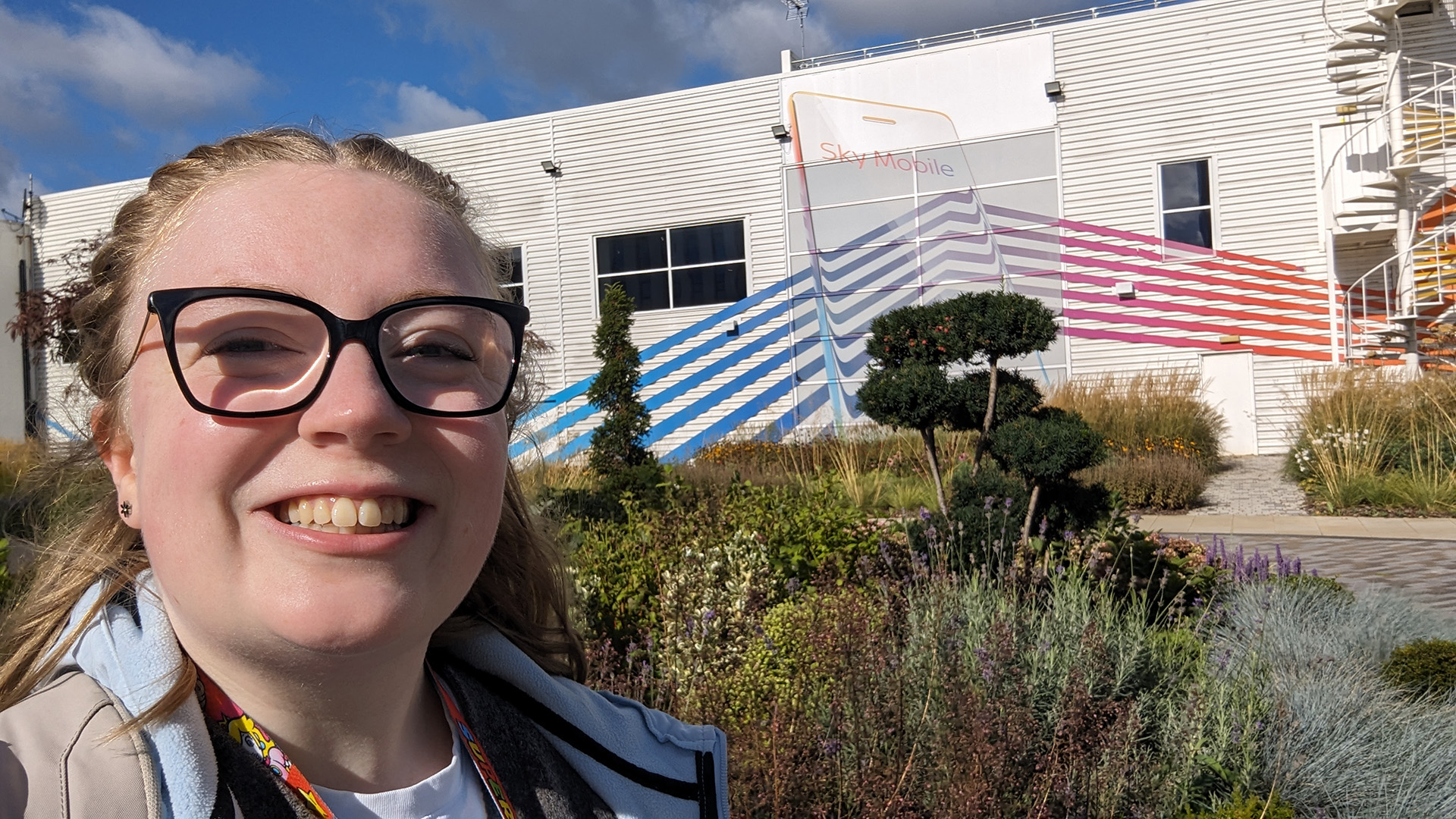 Carys taking selfie onsite at Sky Osterley. Carys is positioned on the left of the screen, showing off the green plants from a nature path just in front of the Sky Mobile building - featuring a spectrum colour shining through a mobile device.