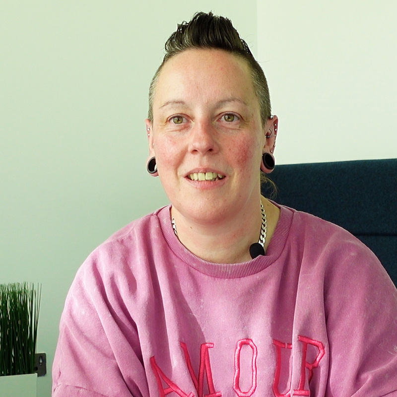 Person with short hair, piercings, and a pink sweatshirt sitting in an office with a small plant in the background.