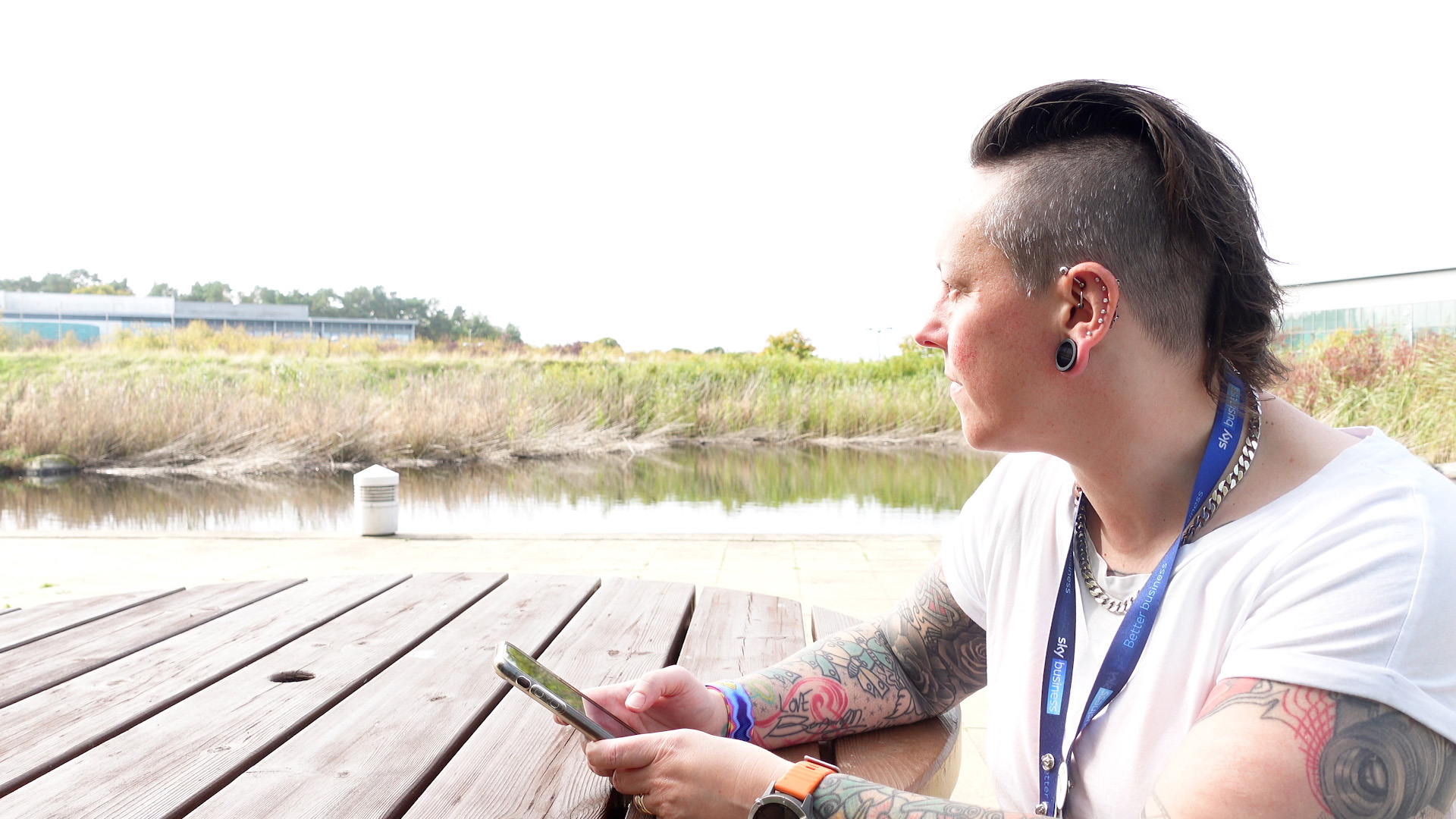 Kirstin sitting on a picnic bench outside the Sky Watermark building. Kirstin is looking into the distance, where the nearby pond, and grassland is featured on the left of the screen. Kirstin is holding her mobile phone.