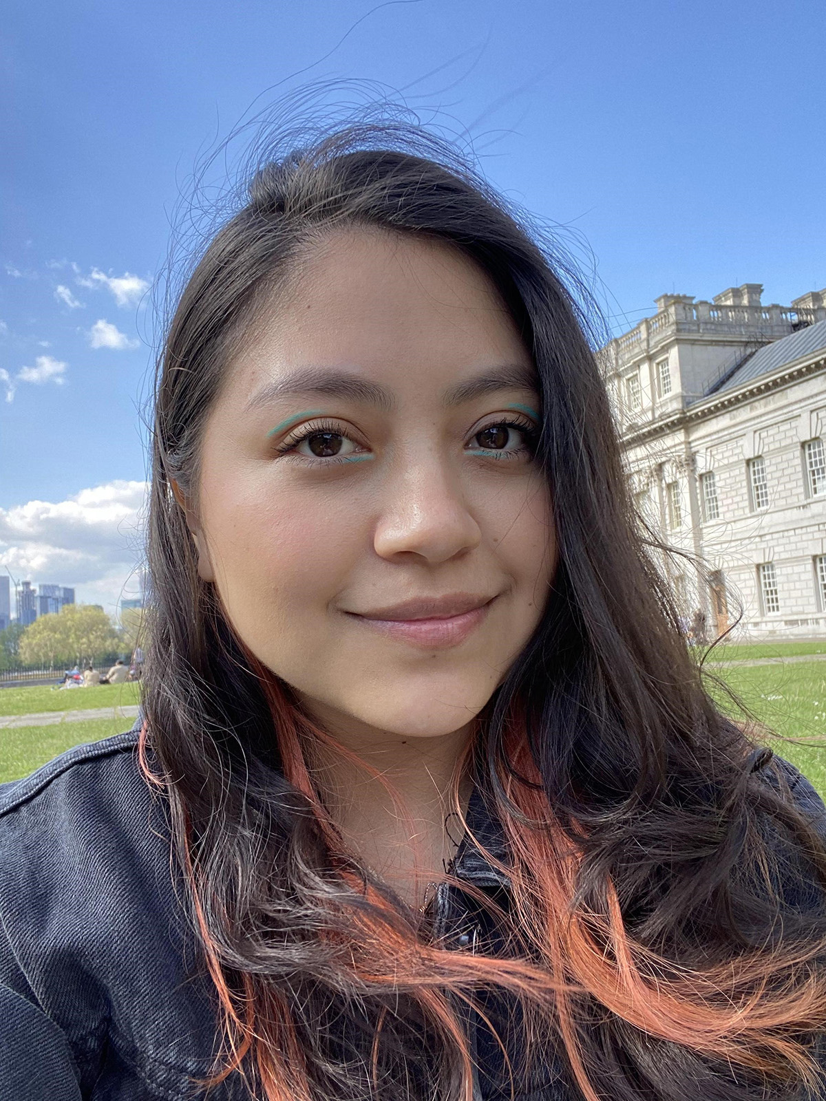 Leslie taking a selfie in front of a backdrop of a stately home building, with a cityscape further in the background.. Leslie looks into camera, as her hair is windswept.
