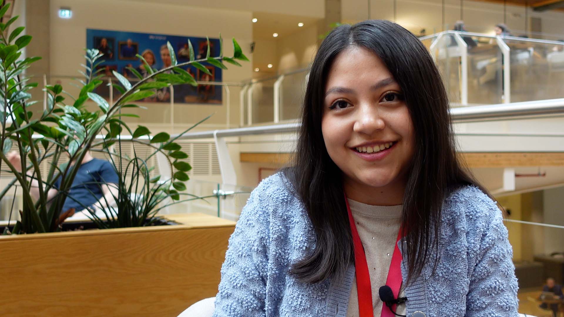 Leslie looks off to the left of the camera in front of a vista of the Sky Osterley office. A potted plant is visible to the left of Leslie, as colleagues have conversations in the background at one of our many on-site cafes.