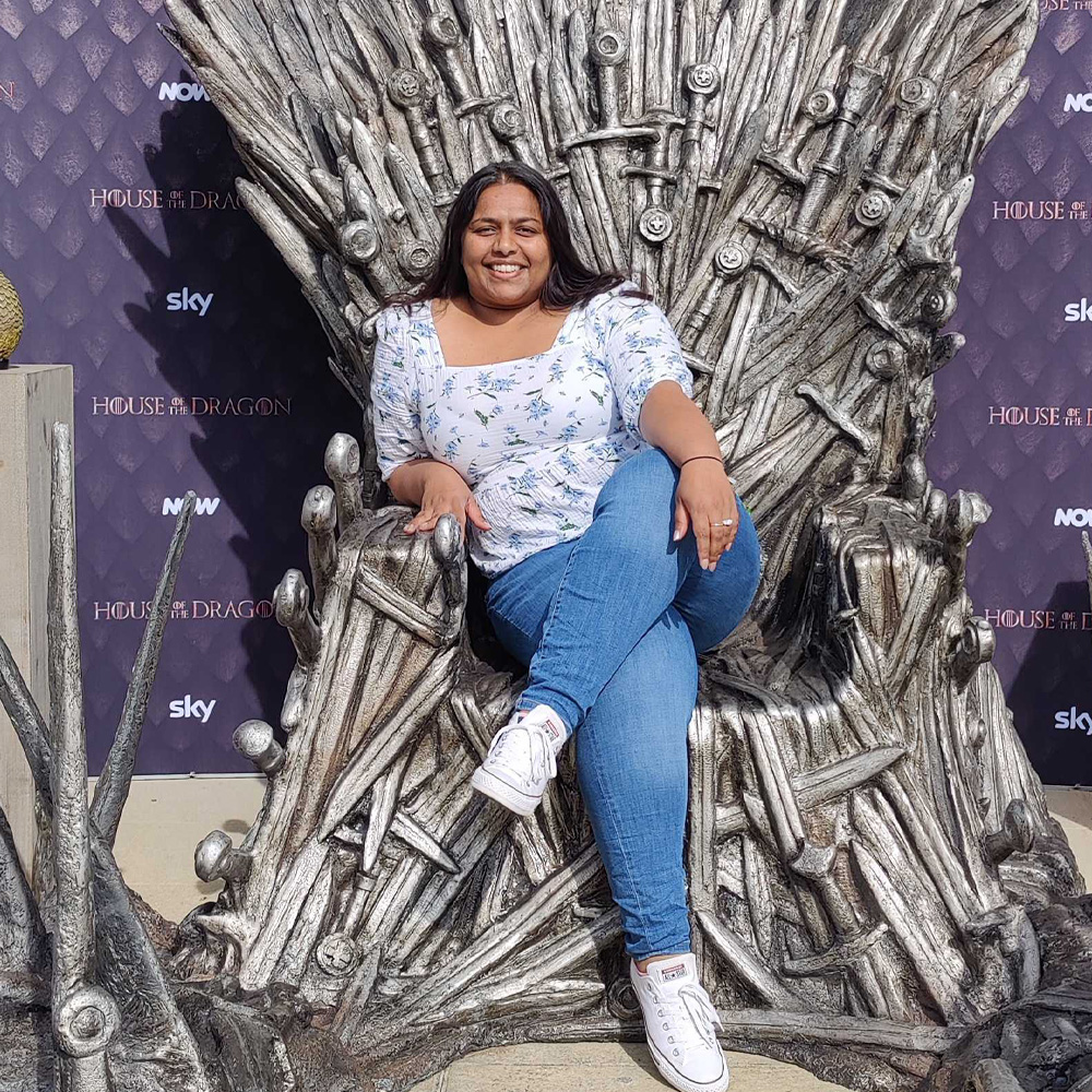 Lina sits cross legged on the iron throne, from the hit show Game of Thrones. In the background is a purple dragon scale patterned print with House of the Dragon, Sky and NOW logos.