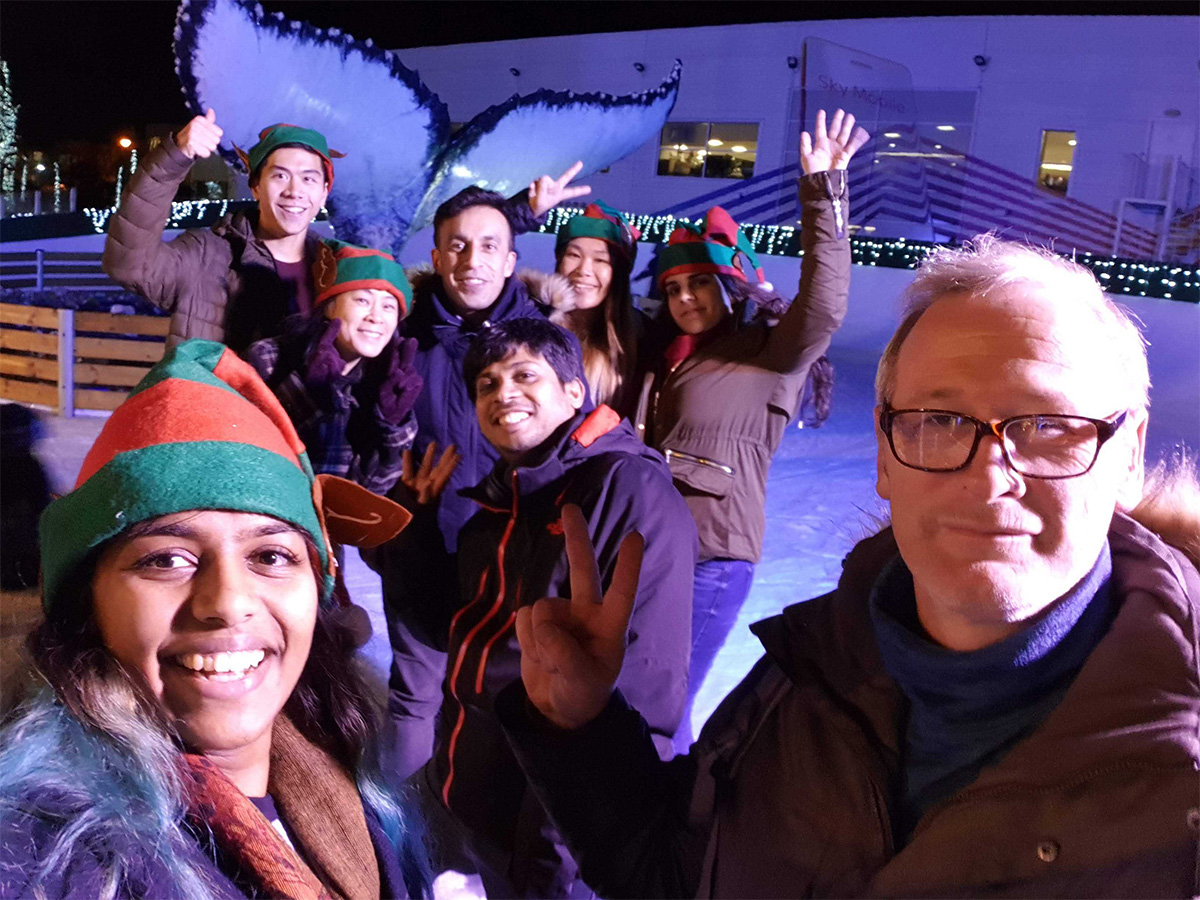 Lina takes a team selfie in front of her team on an ice ink in Sky Osterley. Her team are raising their hands in celebration just in front of the 'Plasticus' whale tail, which in centered in the ice rink.