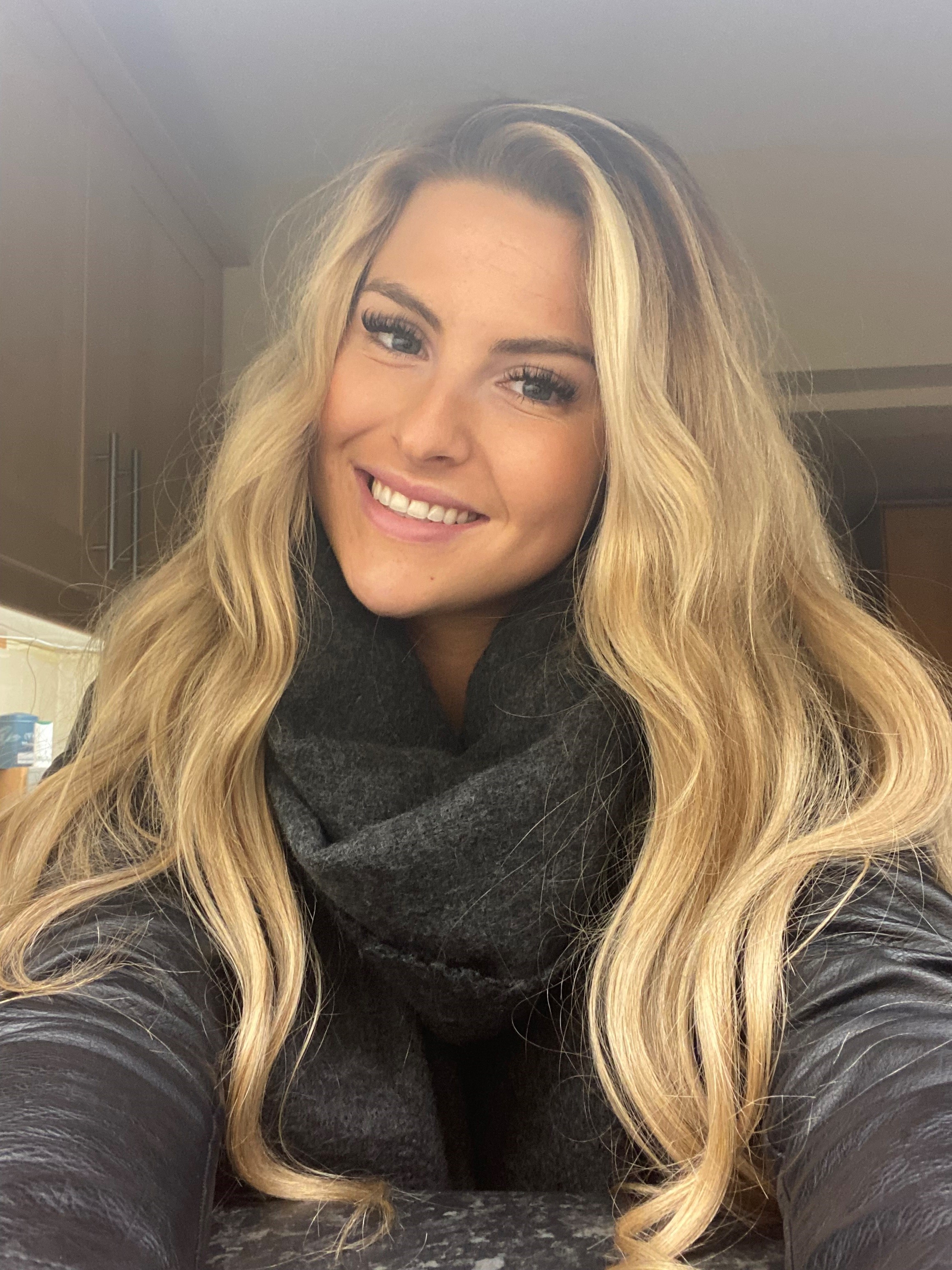 Woman with long blonde hair smiling in a kitchen, wearing a dark scarf and black top.