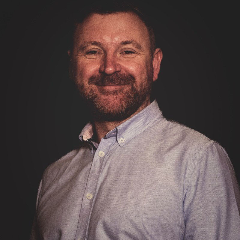 Peter smiles towards the camera, in a blue button shirt. He stands in front of a silhouetted background.