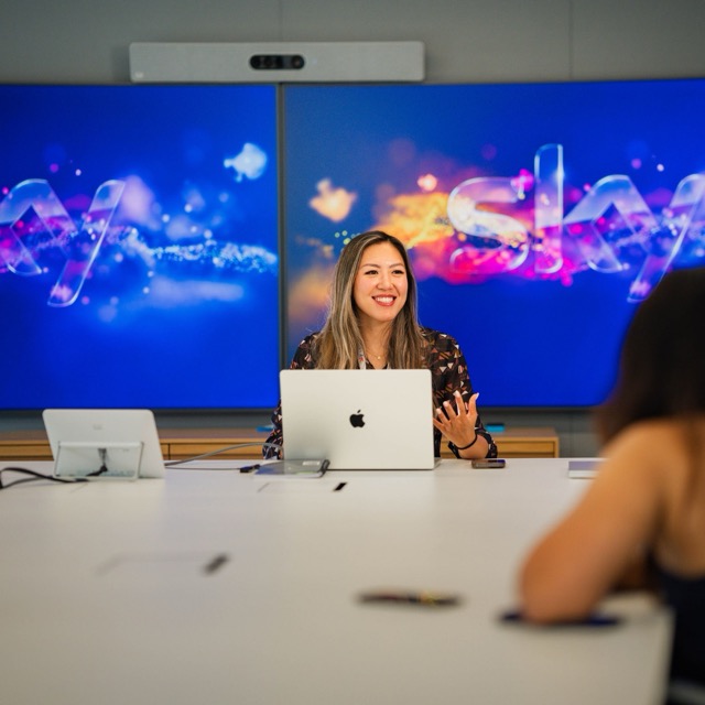 A lady in front of screens, presenting.