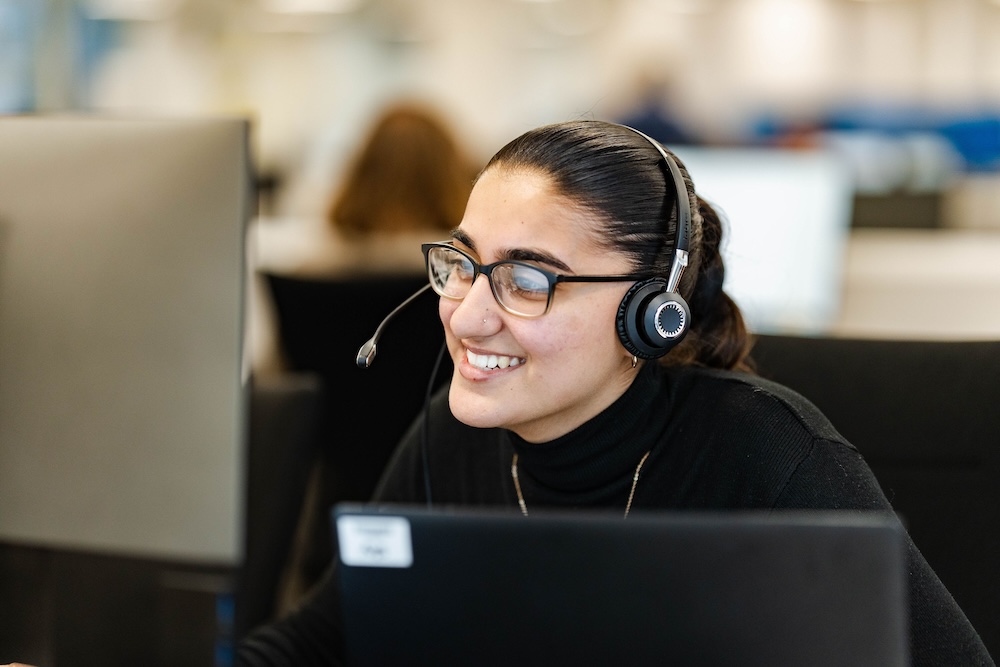 Smiling woman wearing a headset and glasses sits at a desk with a laptop in an office, engaged in a conversation, with blurred coworkers in the background.