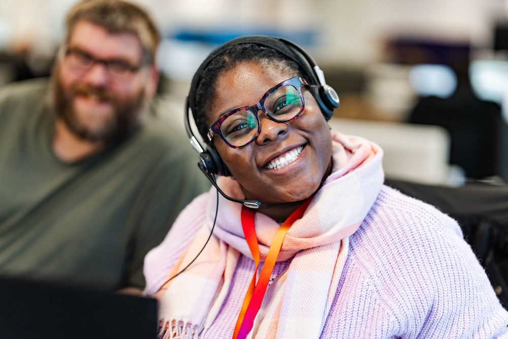 Smiling woman in glasses and headset wearing a pink scarf and lilac sweater sits beside a bearded man in an office, both appearing cheerful and engaged.