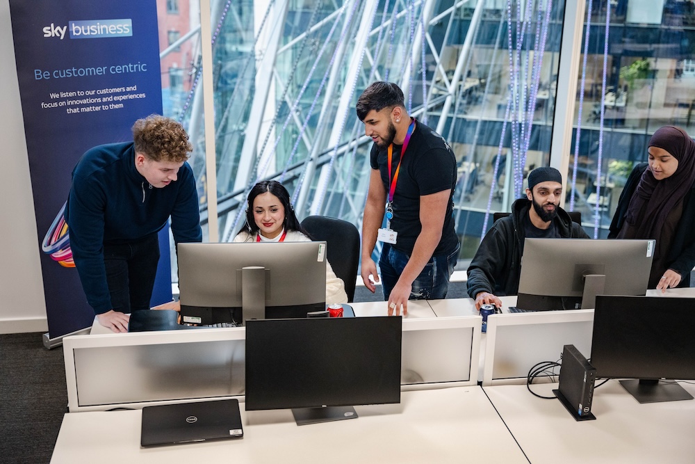Five colleagues gathered around two desks with computers in a modern office, discussing work near a large window and a Sky Business banner.
