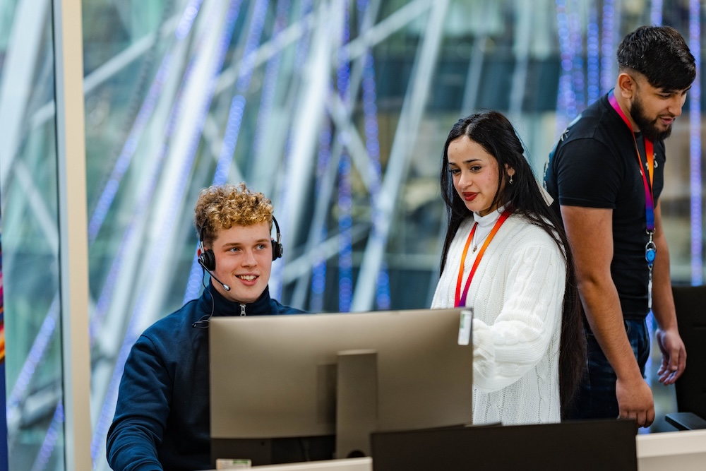 Man wearing a headset sits at a computer smiling while two colleagues stand nearby, all wearing lanyards in a brightly lit office with a glass wall in the background.