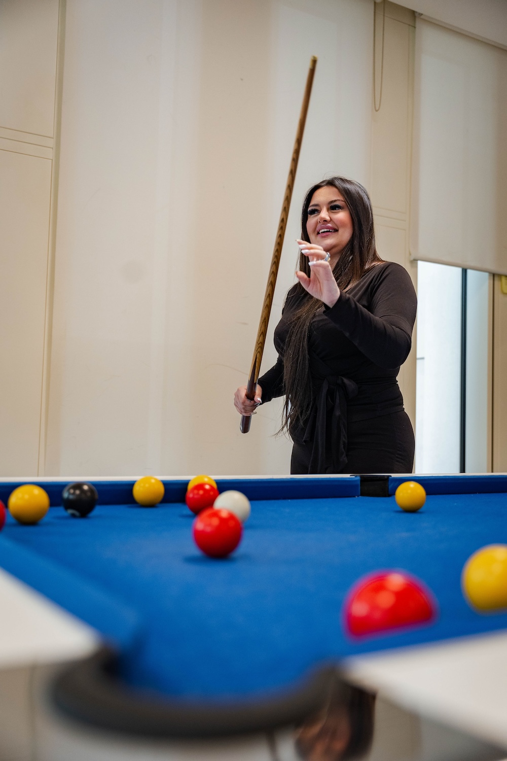 Smiling woman in black outfit holding a cue stick stands near a blue pool table with red, yellow, and black balls scattered across it in a bright indoor space.