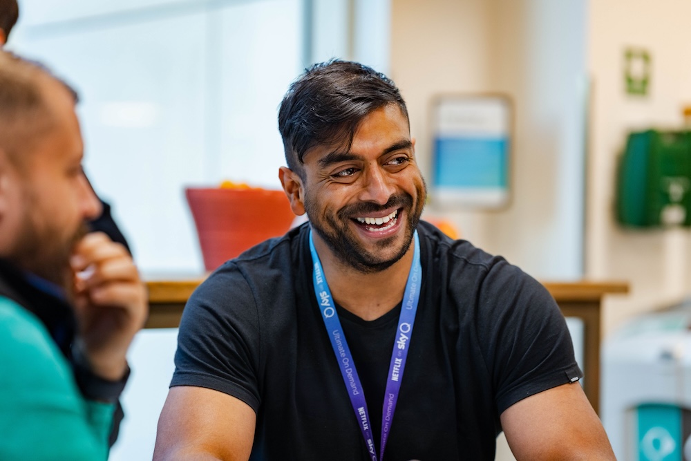 Smiling man wearing a Sky lanyard and black t-shirt sits at a table in a casual office setting, engaged in conversation with a colleague.