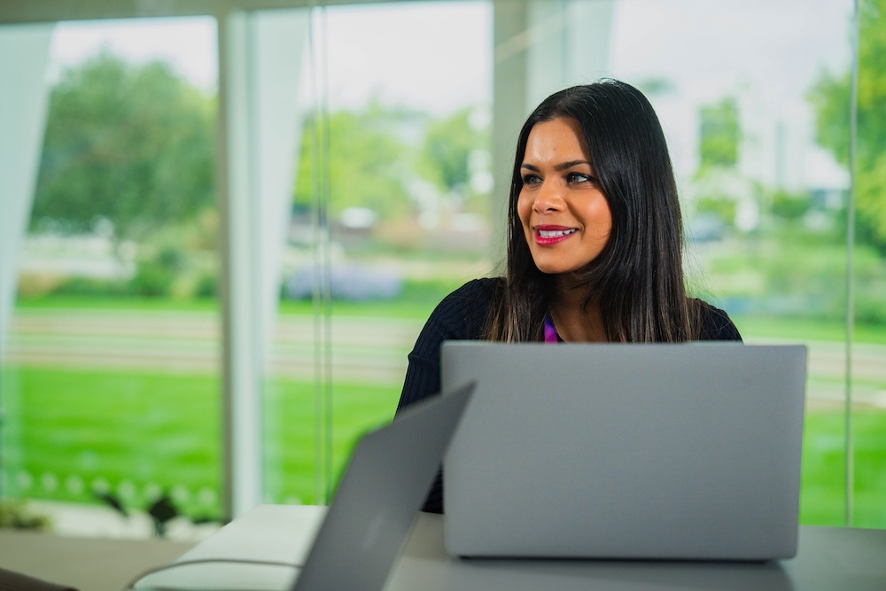 Woman with long dark hair smiles while sitting at a desk with a laptop in a bright office with large windows overlooking a green outdoor space.