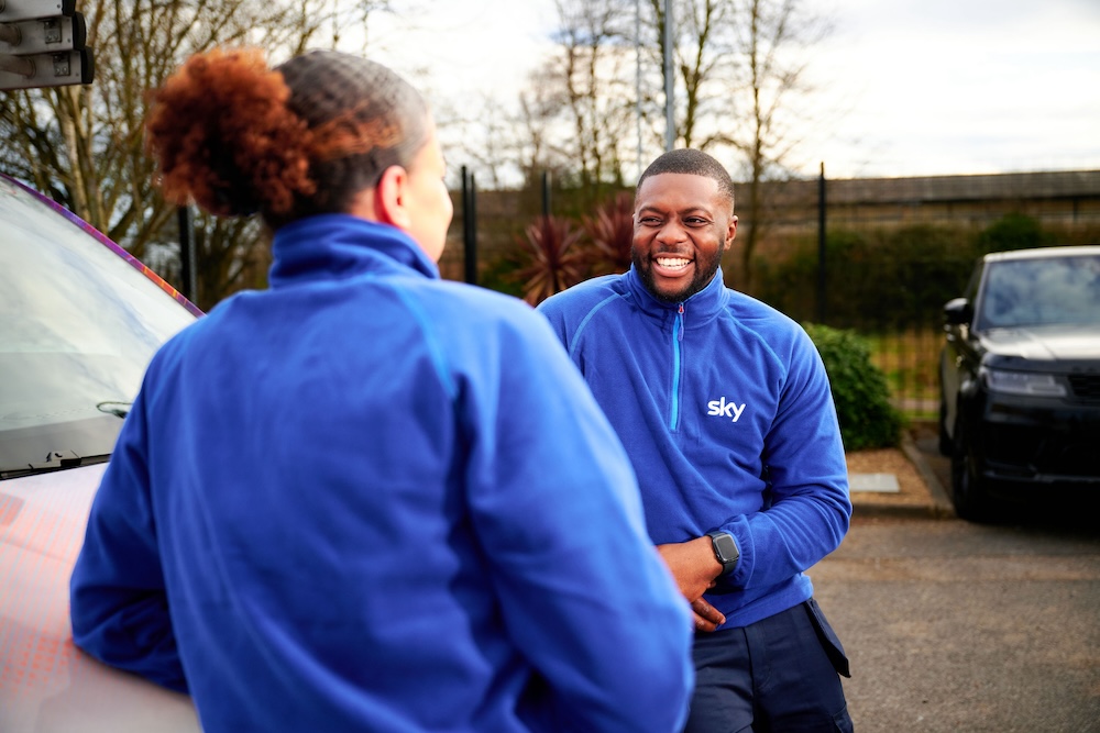 Two Sky employees in blue uniforms chatting beside a branded van in a parking area, both smiling warmly.