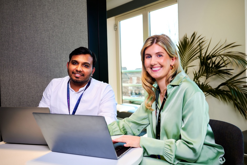 Two colleagues smile at the camera while seated at a desk with laptops in a bright office, with large windows and a leafy indoor plant behind them.