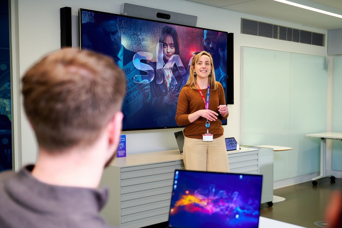 Woman giving a presentation in front of a large screen displaying Sky branding and drama imagery, speaking to seated colleagues with laptops in a modern meeting room.