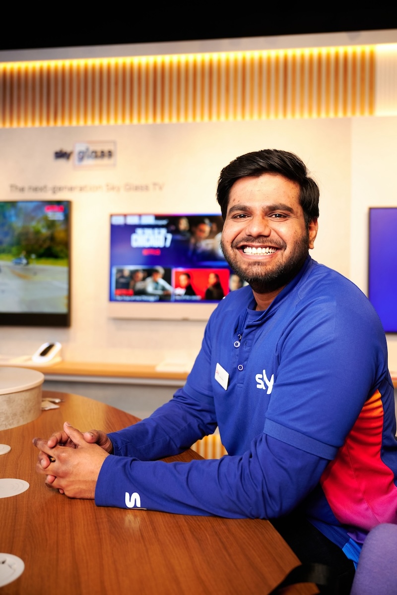 Smiling Sky employee in a blue and pink shirt leans on a wooden counter, with Sky Glass TVs showing vibrant content on the wall behind him.