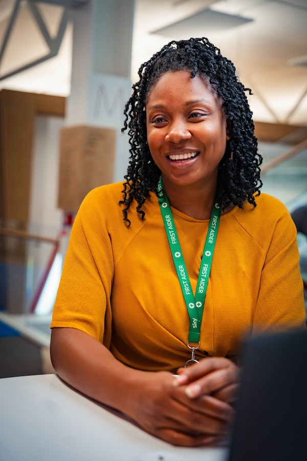 Smiling woman in a mustard top wearing a green “First Aider” lanyard, seated at a table in a bright modern office with blurred background details.