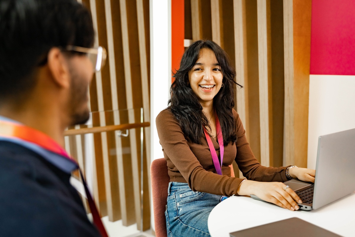 Smiling woman with a laptop sits at a round table wearing a pink lanyard, speaking to a colleague in a modern office with wood panel accents and red walls.