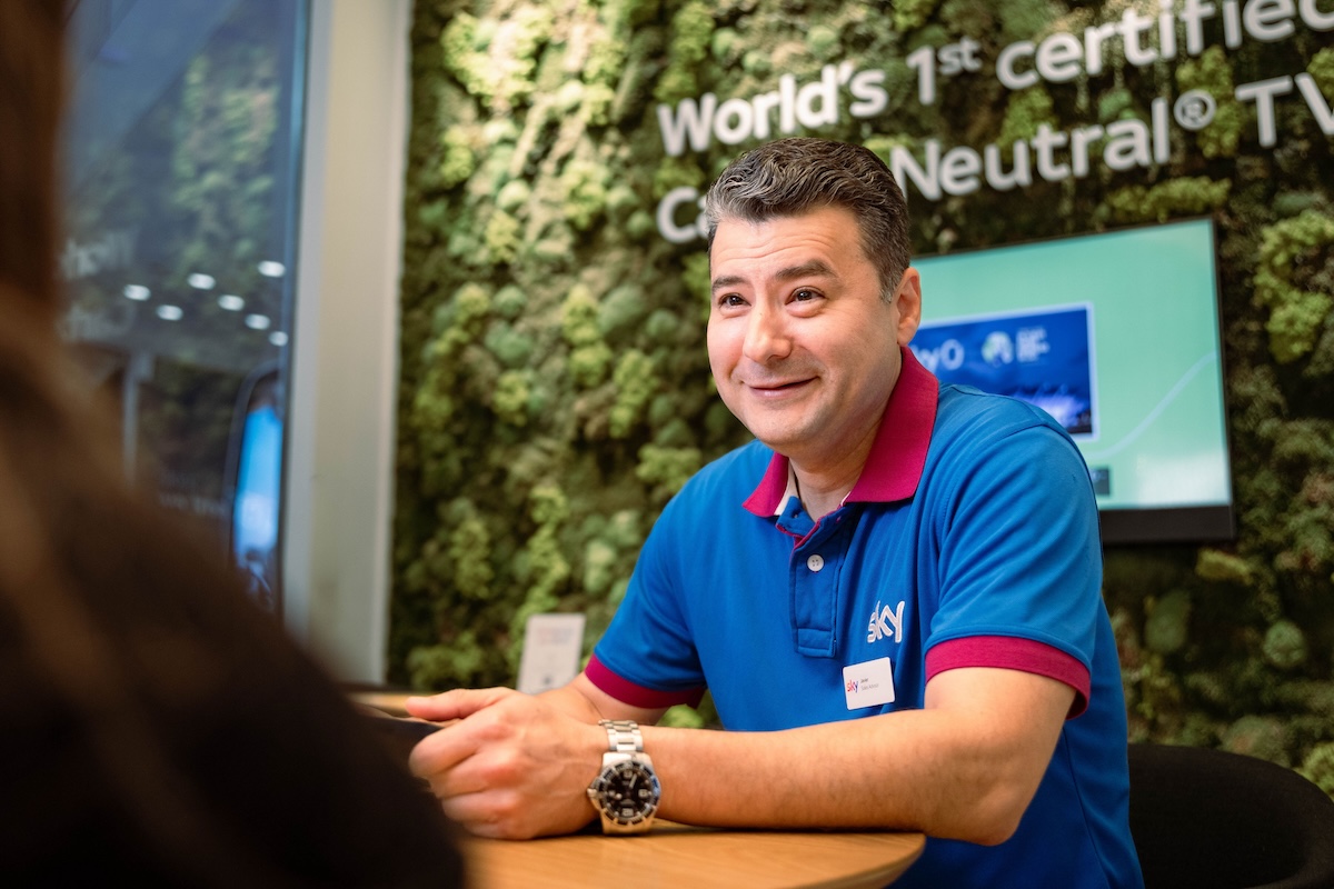 Man in a blue Sky-branded polo shirt smiling at a table, with a watch on his wrist and a green plant wall and Sky signage behind him.